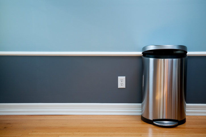 Open trash can in empty room with wood flooring, blue wainscoting and a power outlet.